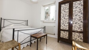 A bedroom with a bed, a small wooden table, a wicker chair, a window with a view, and a wardrobe with floral wallpaper. - House with Big Terrace in Seaside, Syracuse - Ortigia (Siracusa)