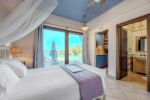 A hotel room with a bed, a view of the sea, a ceiling fan, and a bathroom with a sink and mirror. - Villa Scorpio, Seafront Luxury & Infinity Pool (LAVRIO)