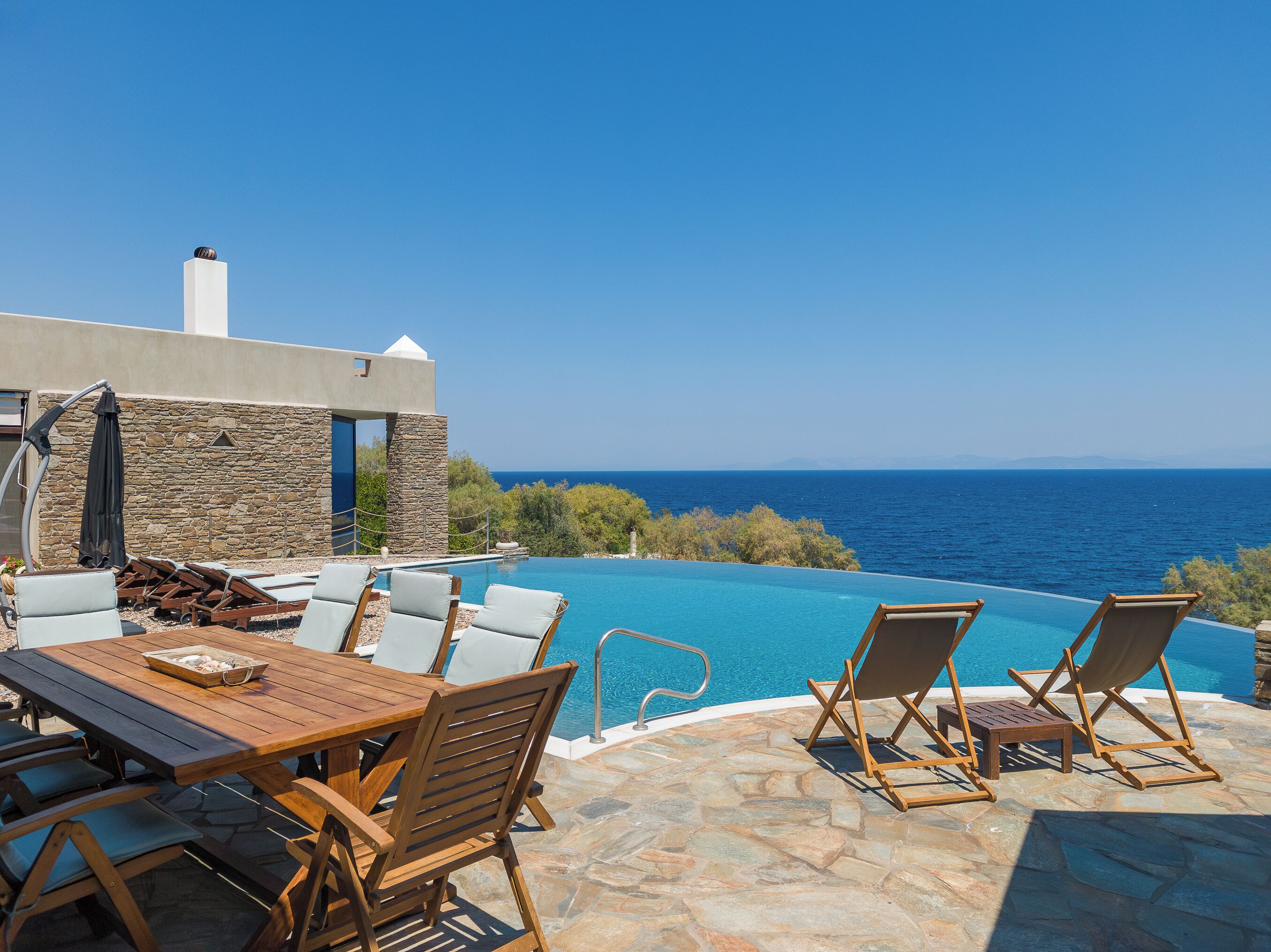 An infinity pool with a clear view of the sea, a wooden table with chairs, and a stone wall.