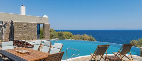 An infinity pool with a clear view of the sea, a wooden table with chairs, and a stone wall.