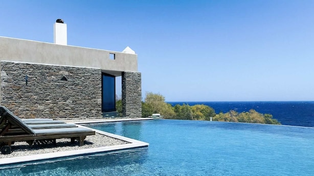 A modern infinity pool with a clear view of the ocean, a stone building with a blue door, and lounge chairs.