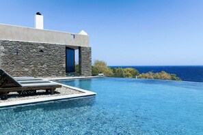 A modern infinity pool with a clear view of the ocean, a stone building with a blue door, and lounge chairs. - Villa Scorpio, Seafront Luxury & Infinity Pool (LAVRIO)