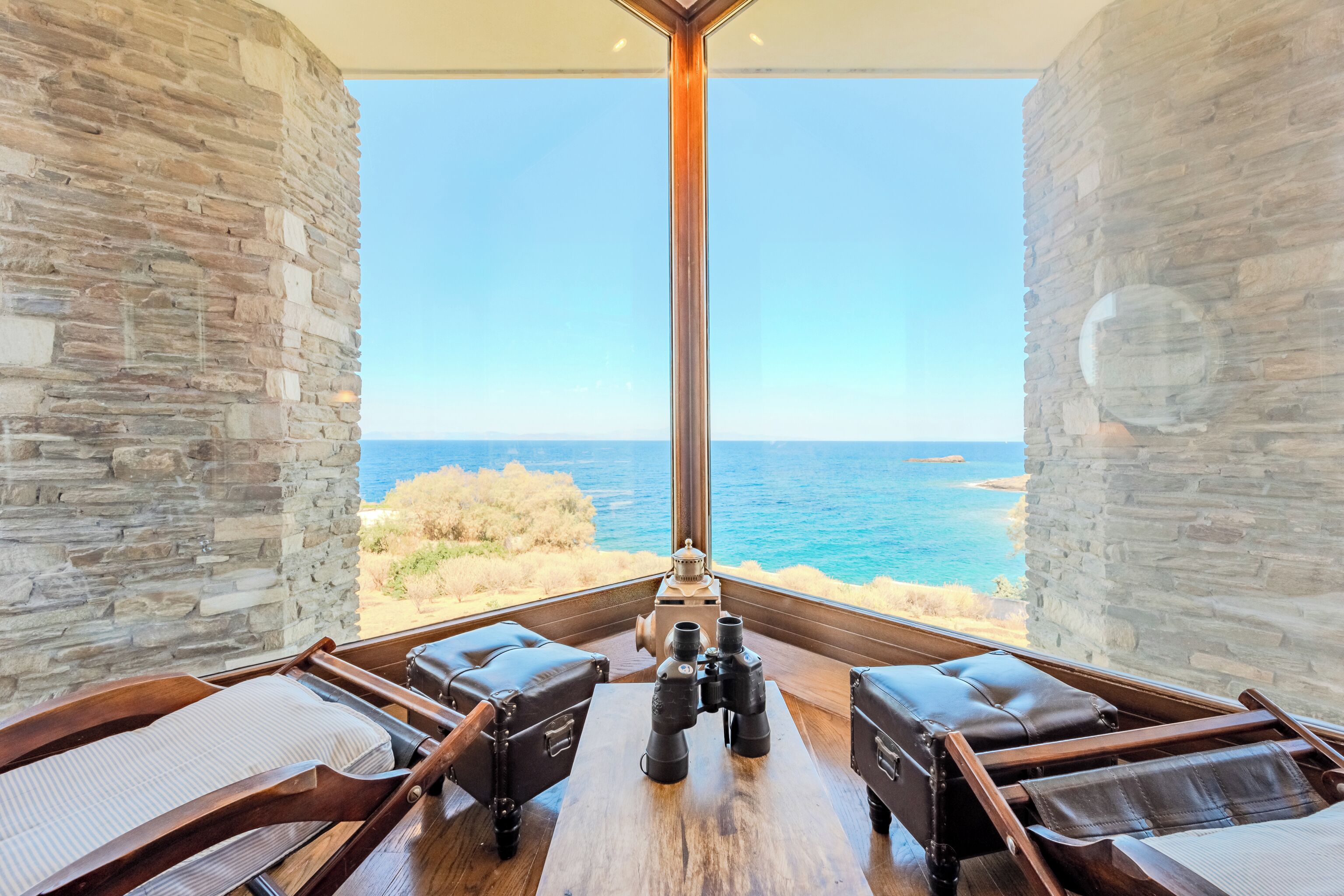 Une pièce avec un mur en pierre et une grande fenêtre donnant sur la mer. On y trouve deux fauteuils en cuir et une table en bois sur laquelle sont posées des jumelles.