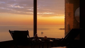 A room with a large window overlooking a sunset over the sea, a chair, and a table with a candle.