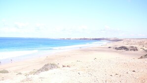 Una amplia playa de arena con un océano azul cristalino, acantilados rocosos a lo lejos y algunas personas nadando.