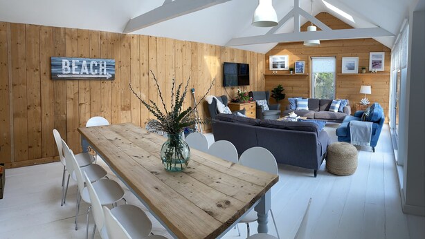 Ein großzügiger Wohnbereich mit einem Holzesstisch, weißen Stühlen, einem grauen Sofa und einem blauen Sofa. Eine Holzwand mit einem 'BEACH'-Schild und einem darüber montierten Fernseher.