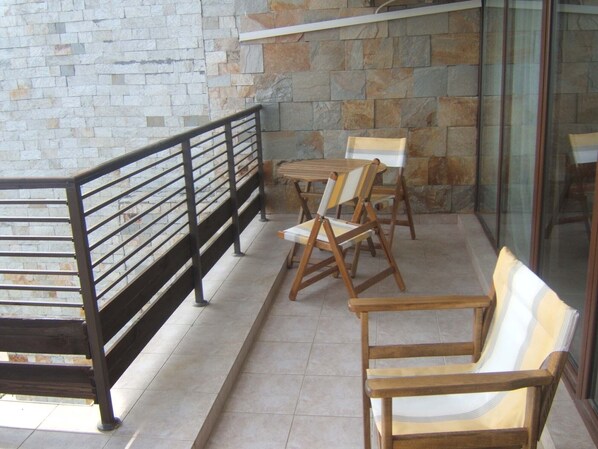 Un balcón con pared de piedra, barandilla de metal y una mesa con sillas.