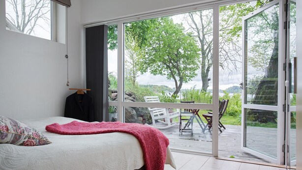 Quarto com cama, manta vermelha, cadeira de madeira e vista para um jardim através de uma porta de vidro deslizante.
