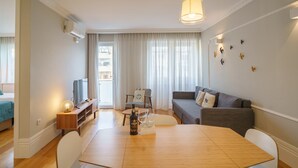 A modern living room with a wooden dining table, a grey sofa, a TV, and a large window with curtains. - Feel Porto Downtown Charming Apartment I (Oporto)