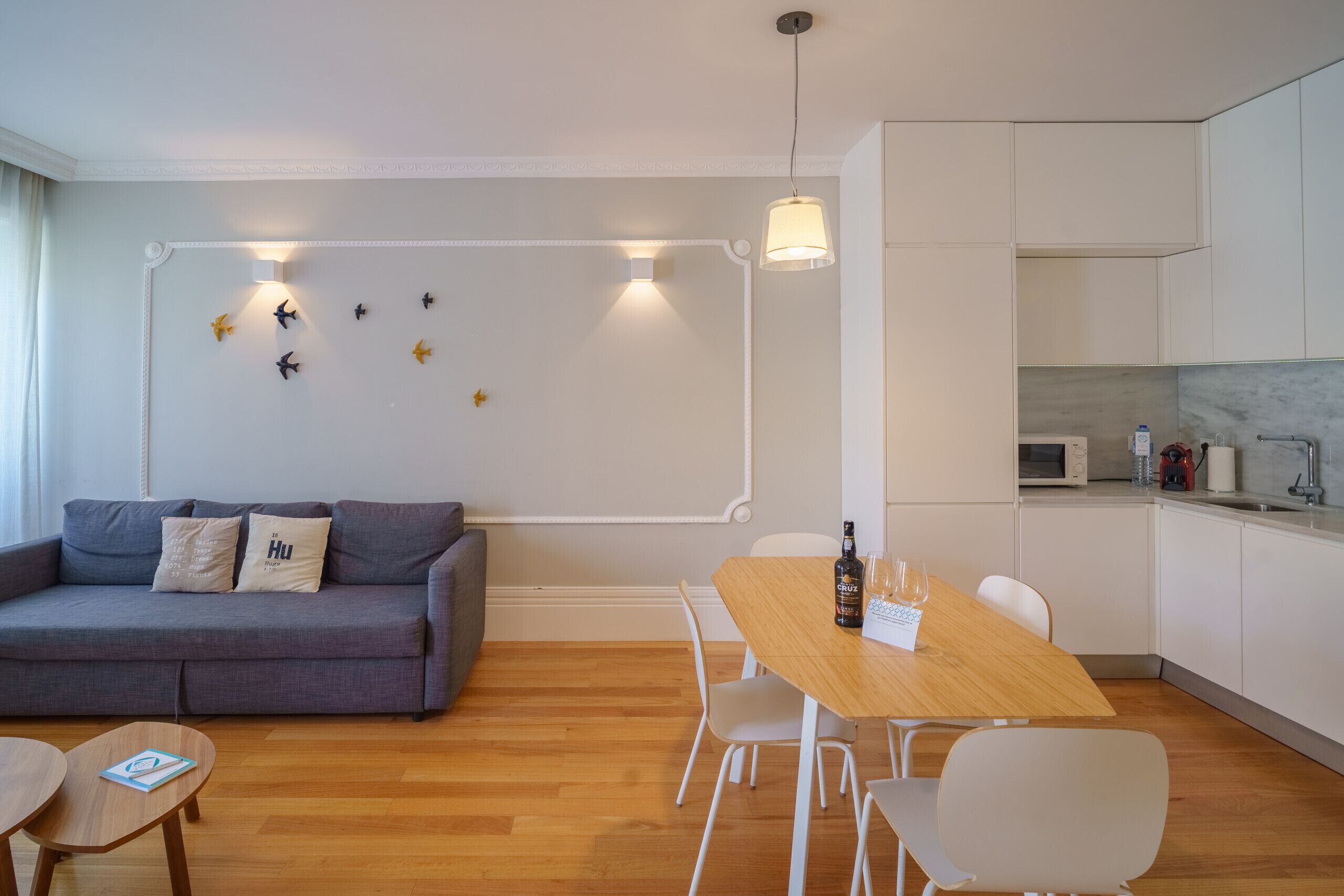 Una sala de estar moderna con un sofá gris, una mesa de comedor de madera y sillas blancas. Hay una zona de cocina con armarios blancos y un microondas.