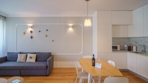 A modern living room with a grey sofa, wooden dining table, and white chairs. There is a kitchen area with white cabinets and a microwave. - Feel Porto Downtown Charming Apartment I (Oporto)