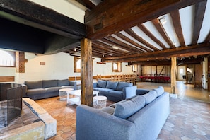 Een ruime kamer met zichtbare houten balken, een grijze bank en een zithoek met tafels en stoelen.