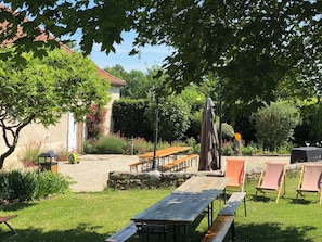 Een tuinpatio met houten tafels en stoelen, omgeven door groen en een stenen muur.