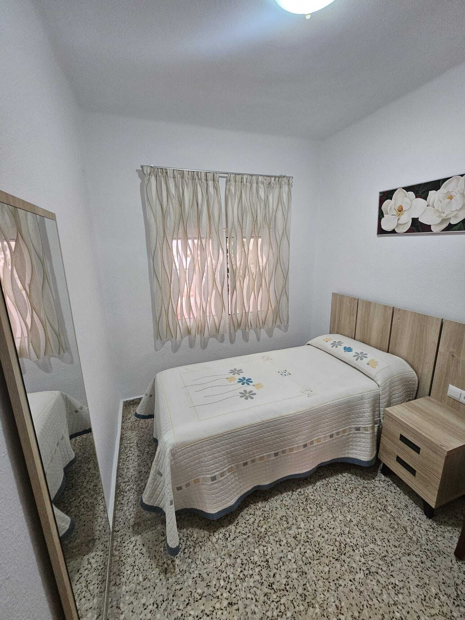 A bedroom with a bed, bedside tables, a mirror, a window with curtains, and a wall-mounted flower painting.