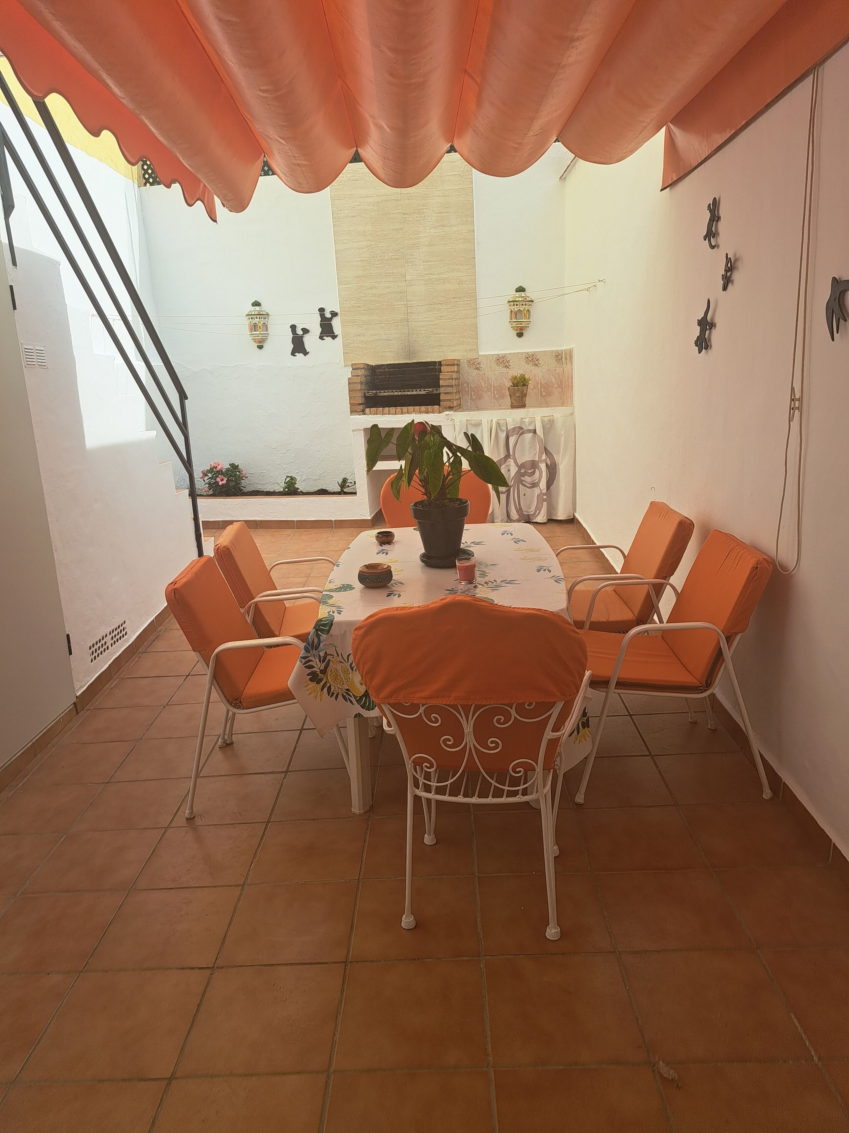 A dining area with a table set for four, surrounded by orange chairs, a potted plant, and a decorative wall hanging.