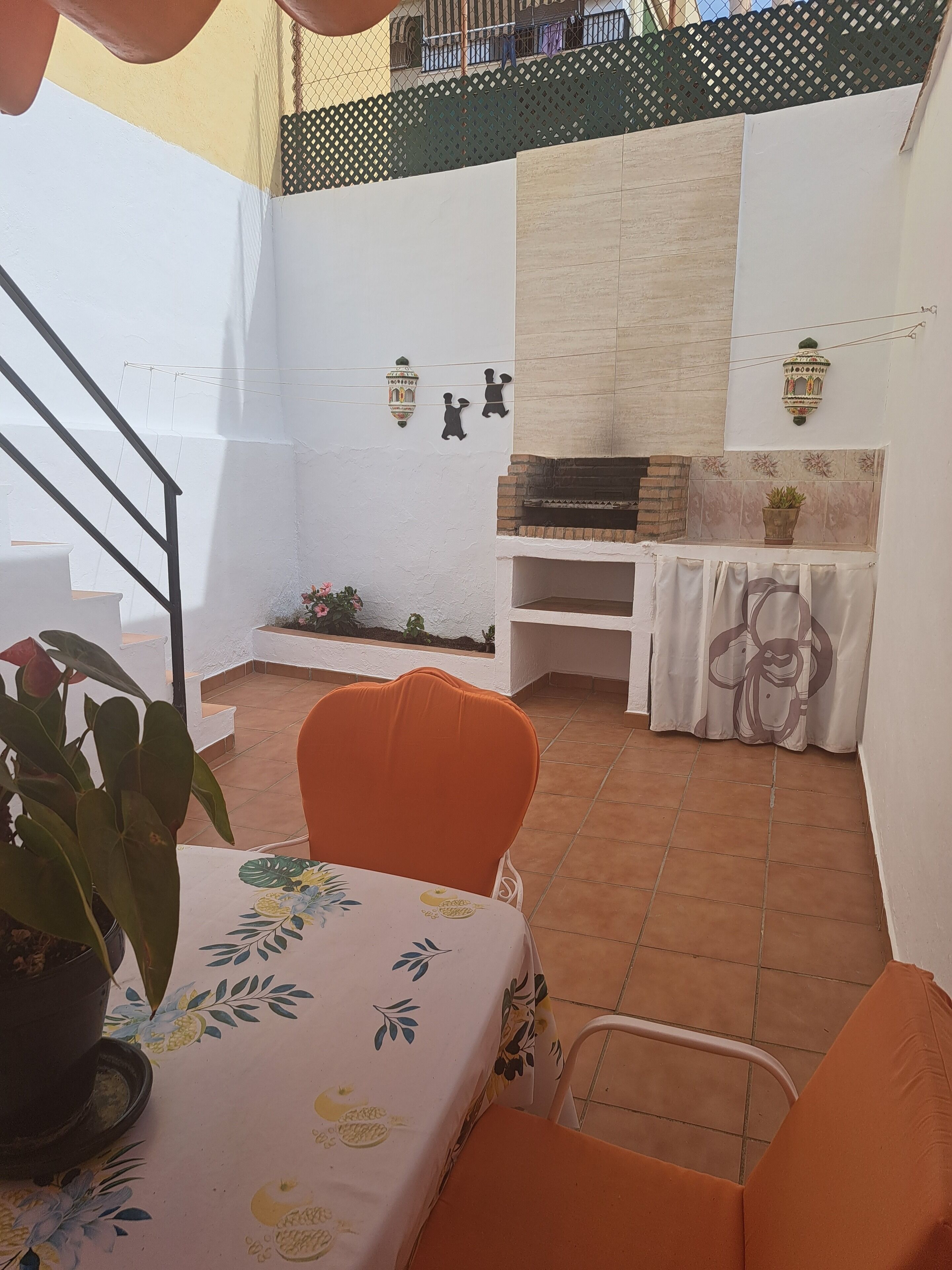Un patio con una pared blanca, un horno de ladrillo, una mesa con mantel floral y sillas naranjas.