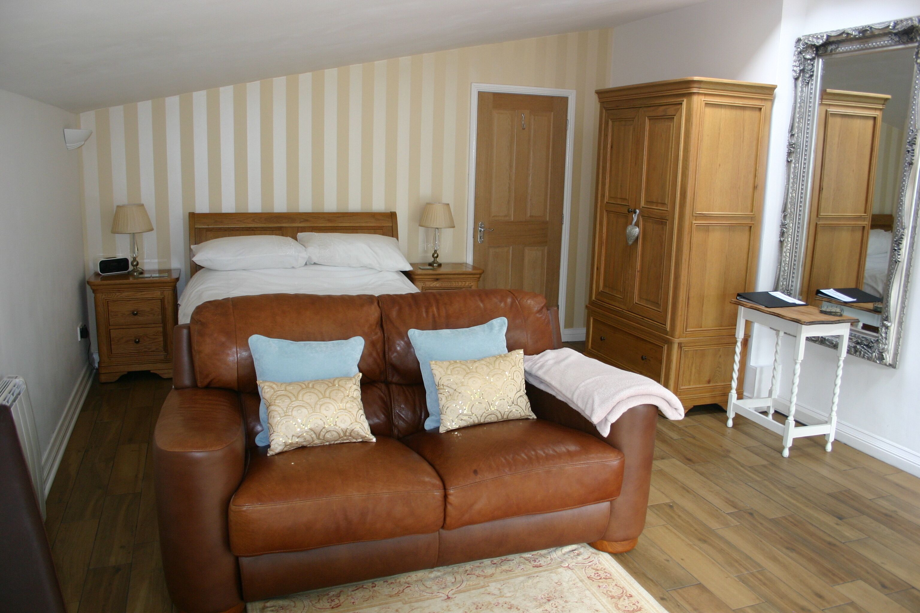A bedroom with a large bed, a brown leather sofa, a wooden wardrobe, and a small table with a lamp.