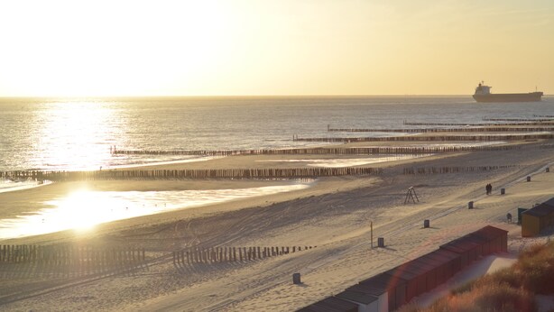 A beach with a pier extending into the water, a ship in the distance, and a sandy area with some structures.
