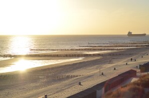 Une plage avec une jetée qui s’avance dans l’eau, un navire à l’horizon et une zone sablonneuse avec quelques constructions.