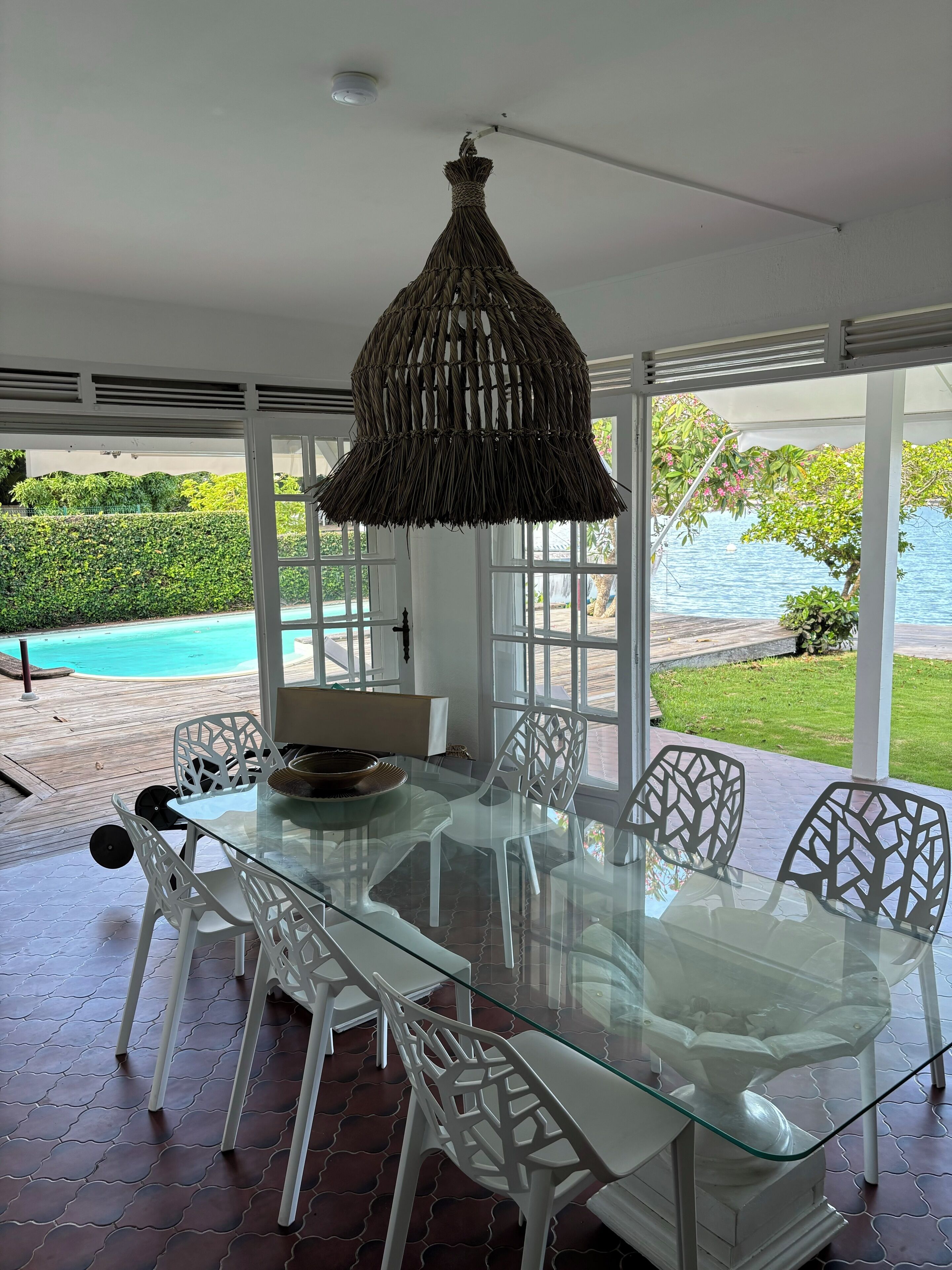 A glass-top dining table with white chairs, a woven pendant light, and a view of a pool and greenery outside.