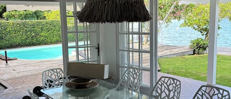 A glass-top dining table with white chairs, a woven pendant light, and a view of a pool and greenery outside.