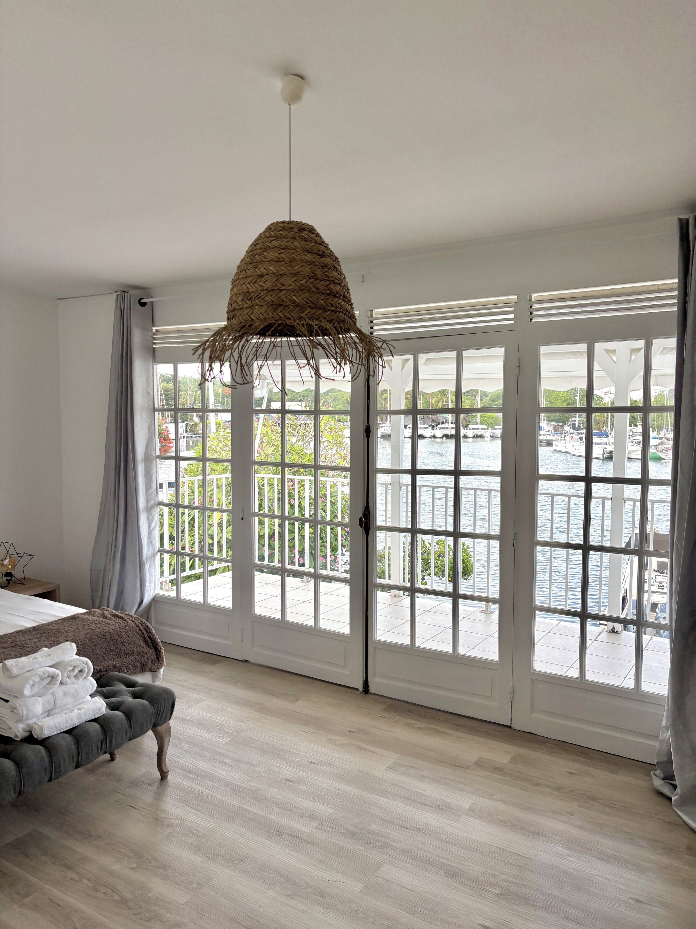 A room with a bed, a bench, a hanging light fixture, and large windows with a view of a marina.