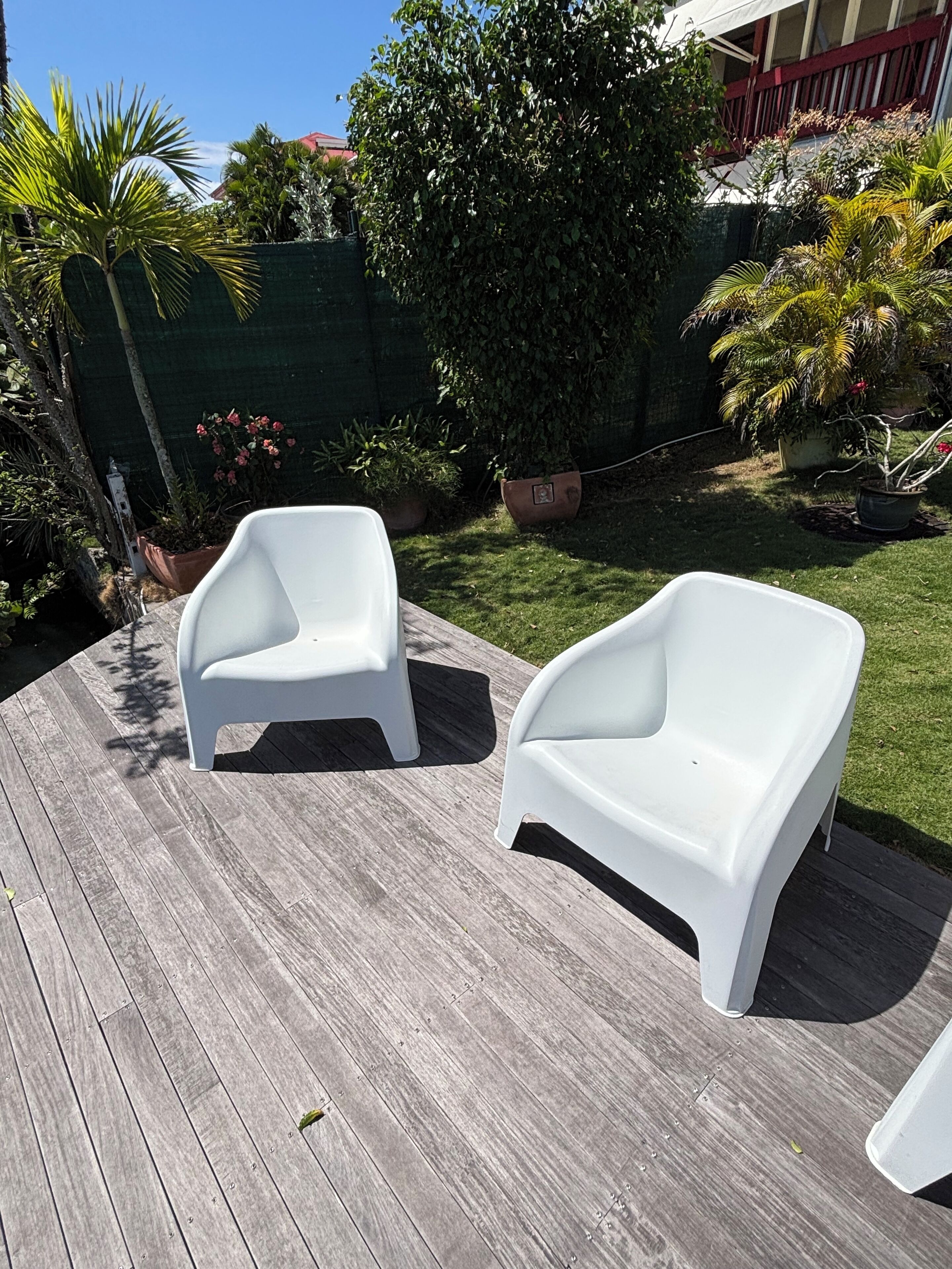 Two white plastic chairs on a wooden deck with a green fence and plants in the background.