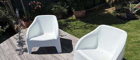 Two white plastic chairs on a wooden deck with a green fence and plants in the background.