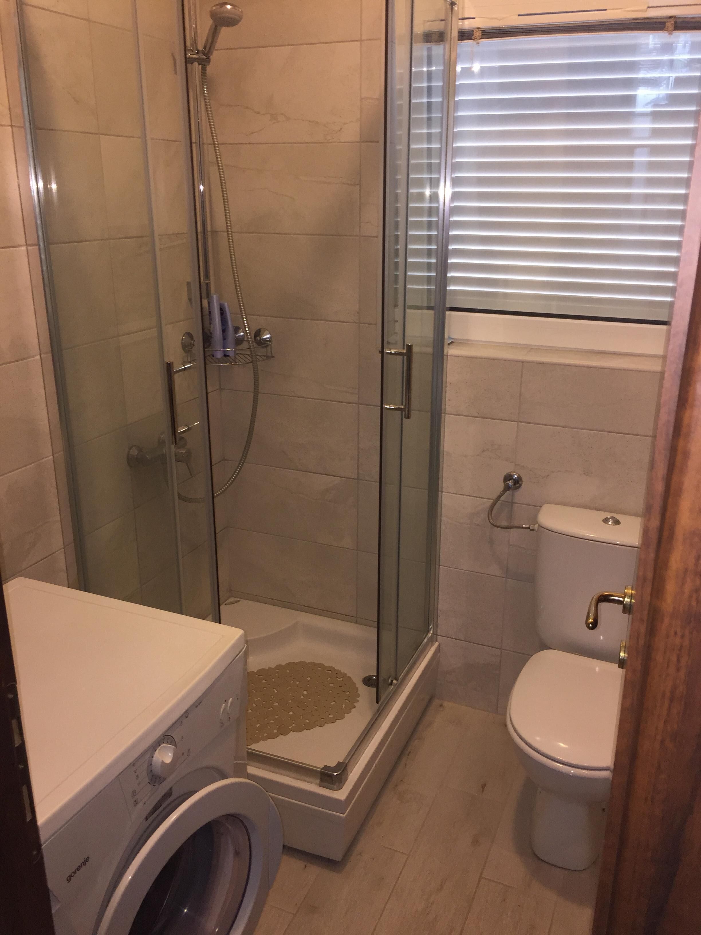 A compact bathroom with a glass-enclosed shower, a washing machine, and a toilet.
