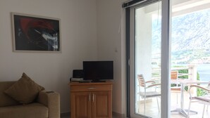 A living room with a sofa, a television on a wooden cabinet, and a sliding glass door leading to an outdoor balcony with chairs. - Ground Floor. Private Terrace, Shared Pool, Views across Fjord to Kotor Old Town (Kotor)