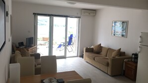 A modern living room with a sofa, a dining table, and a TV. There is a sliding glass door leading to an outdoor area with a chair and a table. - Ground Floor. Private Terrace, Shared Pool, Views across Fjord to Kotor Old Town (Kotor)