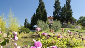 Un giardino con fiori rosa in fiore e uno stagno, circondato da alberi e una casa sullo sfondo.