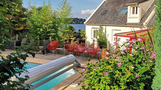 Un espace piscine avec un toboggan, entouré de verdure et de chaises rouges, offrant une vue sur la mer et une maison en arrière-plan.