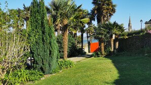 Ein gepflegter Garten mit Palmen, einer Steinmauer und Blick auf ein historisches Gebäude im Hintergrund.