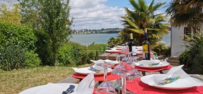 Mesa de comedor con manteles individuales rojos, copas de vino y platos, con vista a un lago y casas a lo lejos.