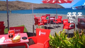 Mobiliario exterior con sillas y sombrillas rojas con la marca Coca-Cola, una mesa con una cesta y una botella, y vista al mar y colinas al fondo.