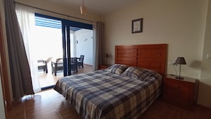 Un dormitorio con cama, mesita de noche, lámpara, silla y una mesa con una computadora portátil.