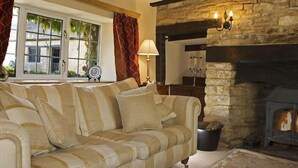 A cozy living room with a striped sofa, a stone fireplace, and a window with curtains. - The Cotswold Manor Hall, Exclusive Hot Tub, Games Barn, 70 acres of Parkland (Bampton)