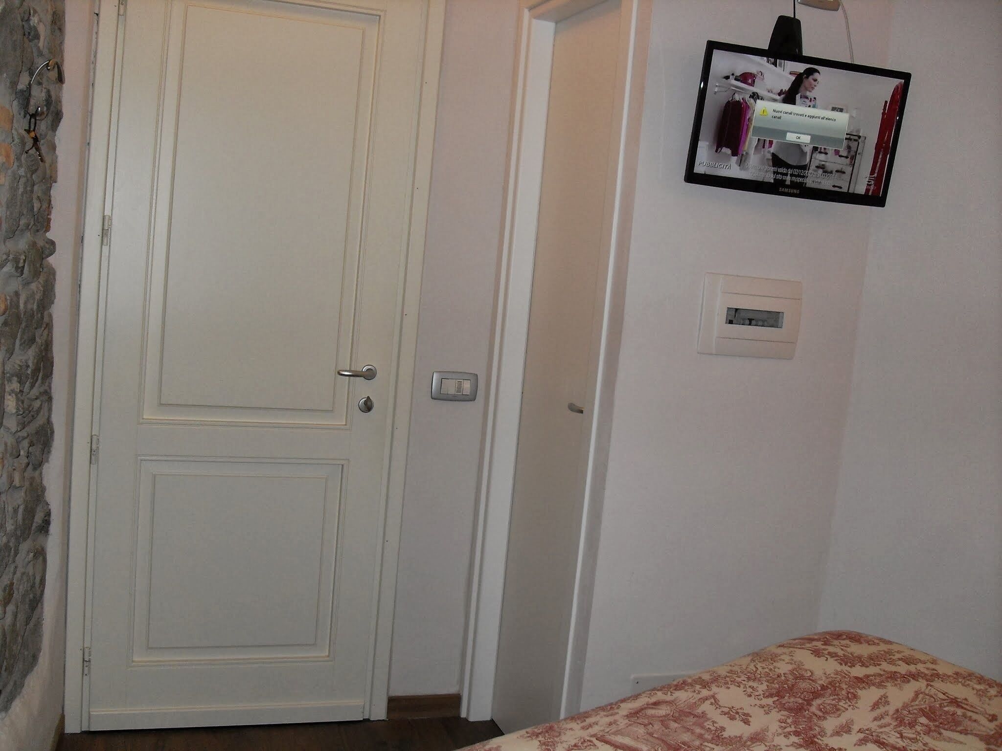 Une pièce avec une porte blanche fermée, un lit recouvert d’une couvre-lit à motifs floraux, un téléviseur fixé au mur et un thermostat.