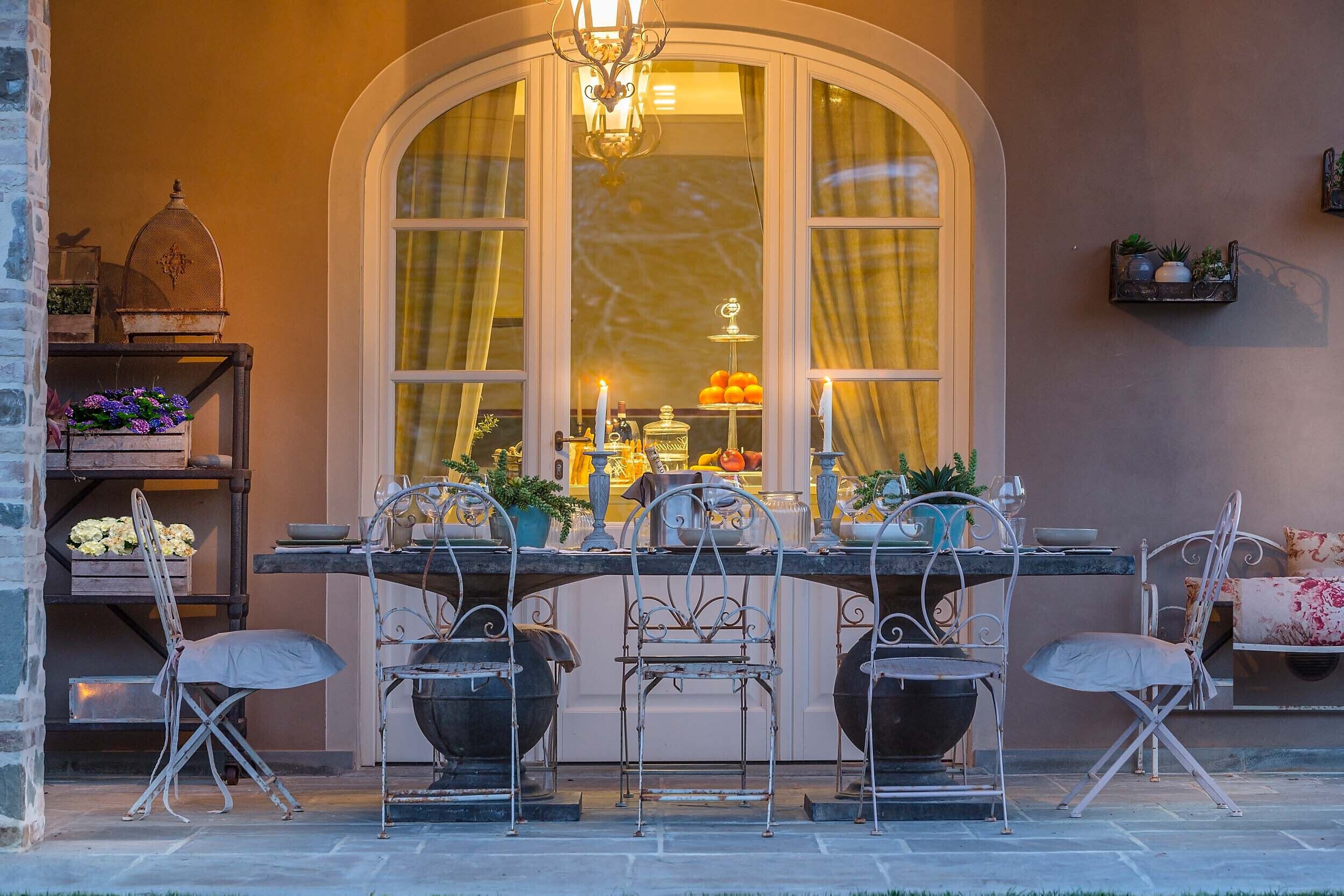 An outdoor dining area with a table and chairs, a chandelier, and a view through a large arched window.