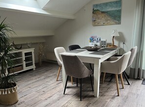 Een eetgedeelte met een witte tafel, grijze stoelen, een bloempot, een schilderij en een radiator.