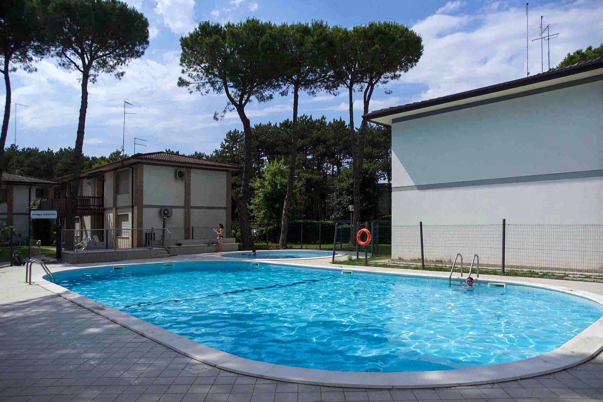 Una piscina con cubierta de baldosas, un puesto de socorrista y un edificio con balcón al fondo.