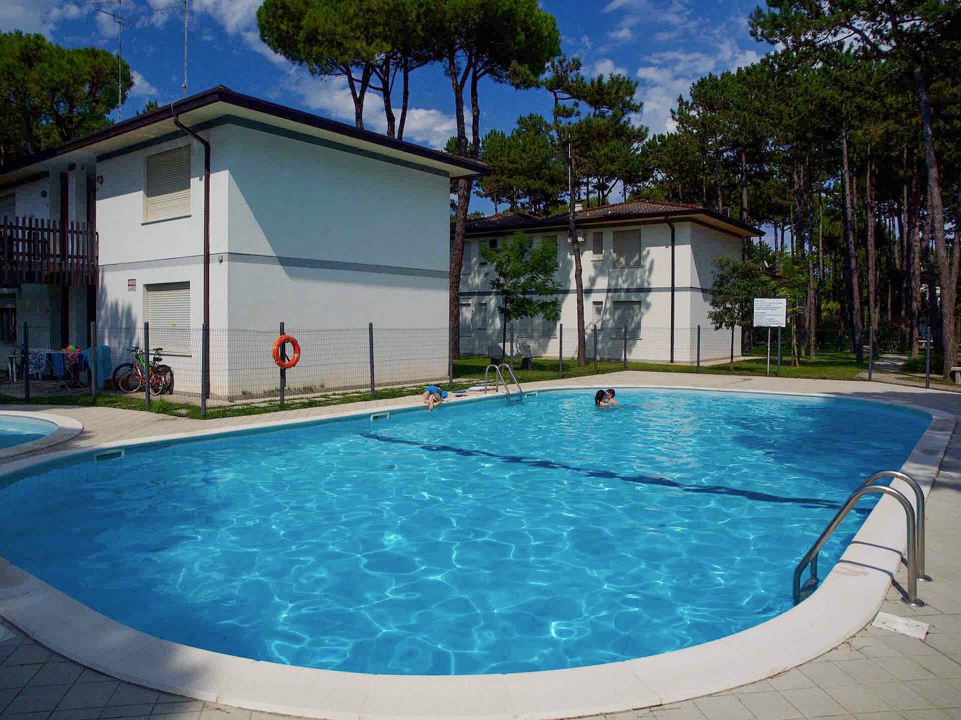 Una piscina con agua cristalina azul, rodeada por una valla y contigua a un edificio de fachada blanca.