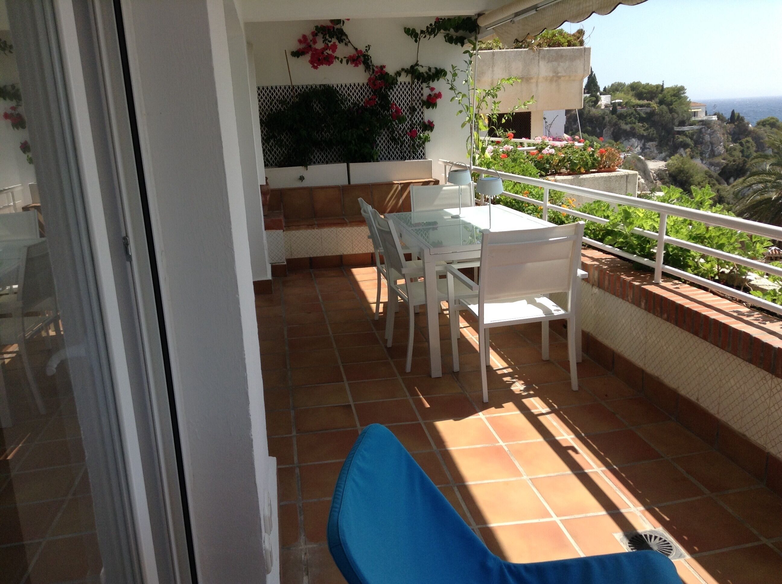 A balcony with a white table and chairs, a blue cushion, and a view of the sea and greenery.