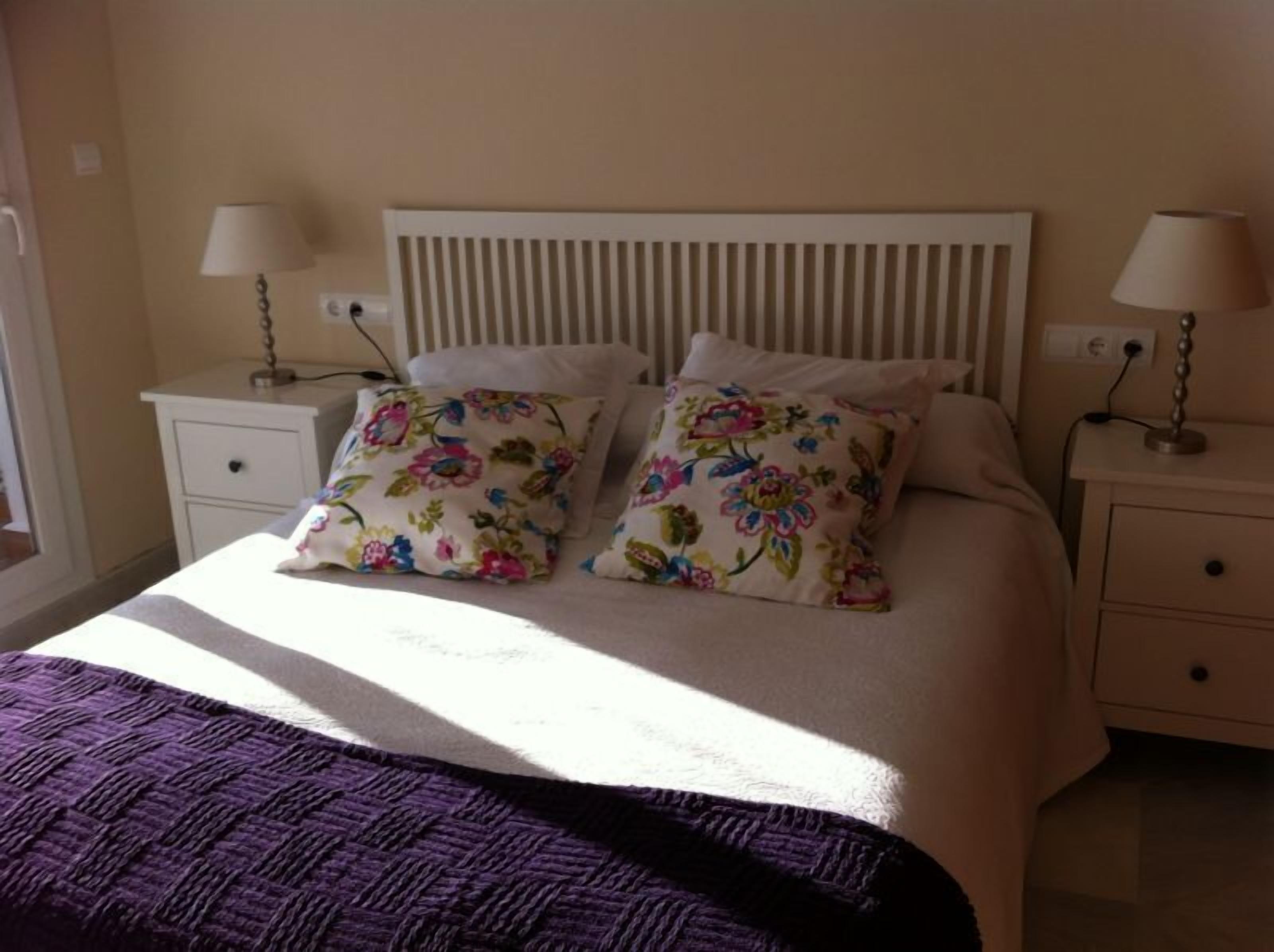 A bed with a white headboard, two floral pillows, a purple blanket, and two bedside tables with lamps.