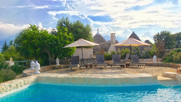 A poolside area with lounge chairs and umbrellas, surrounded by stone walls and greenery.