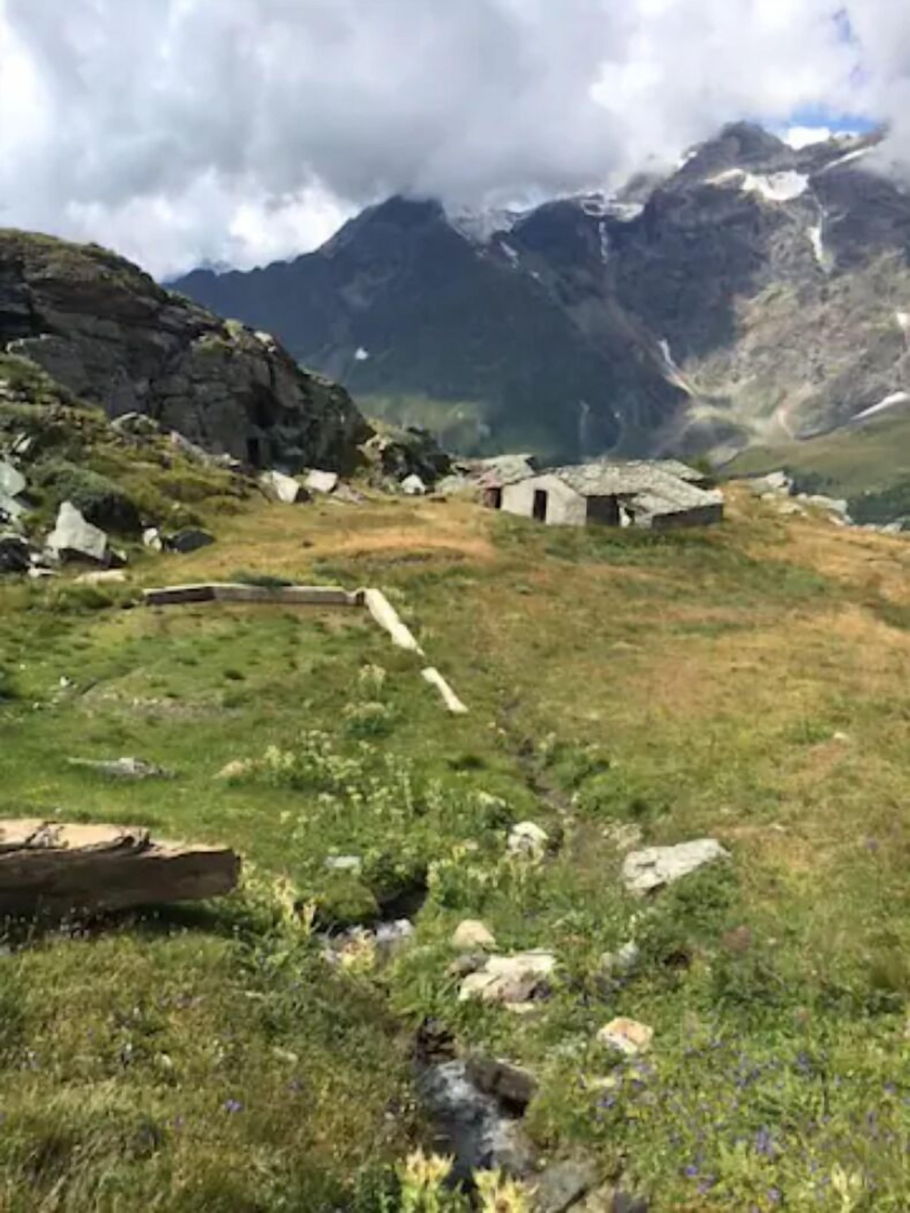 Paesaggio montano rurale con un piccolo gruppo di edifici in pietra, un ruscello e montagne innevate sullo sfondo.