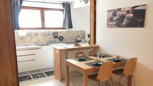 A compact kitchen with a dining area, featuring a wooden table and chairs, a sink, and a window with curtains. - Casa Cervinia Apartment  (Breuil Cervinia)