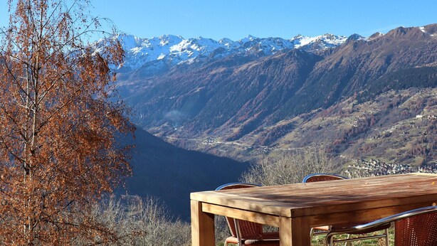 Une table et des chaises en bois, installées sur une colline, offrent une vue sur un paysage montagneux aux sommets enneigés.