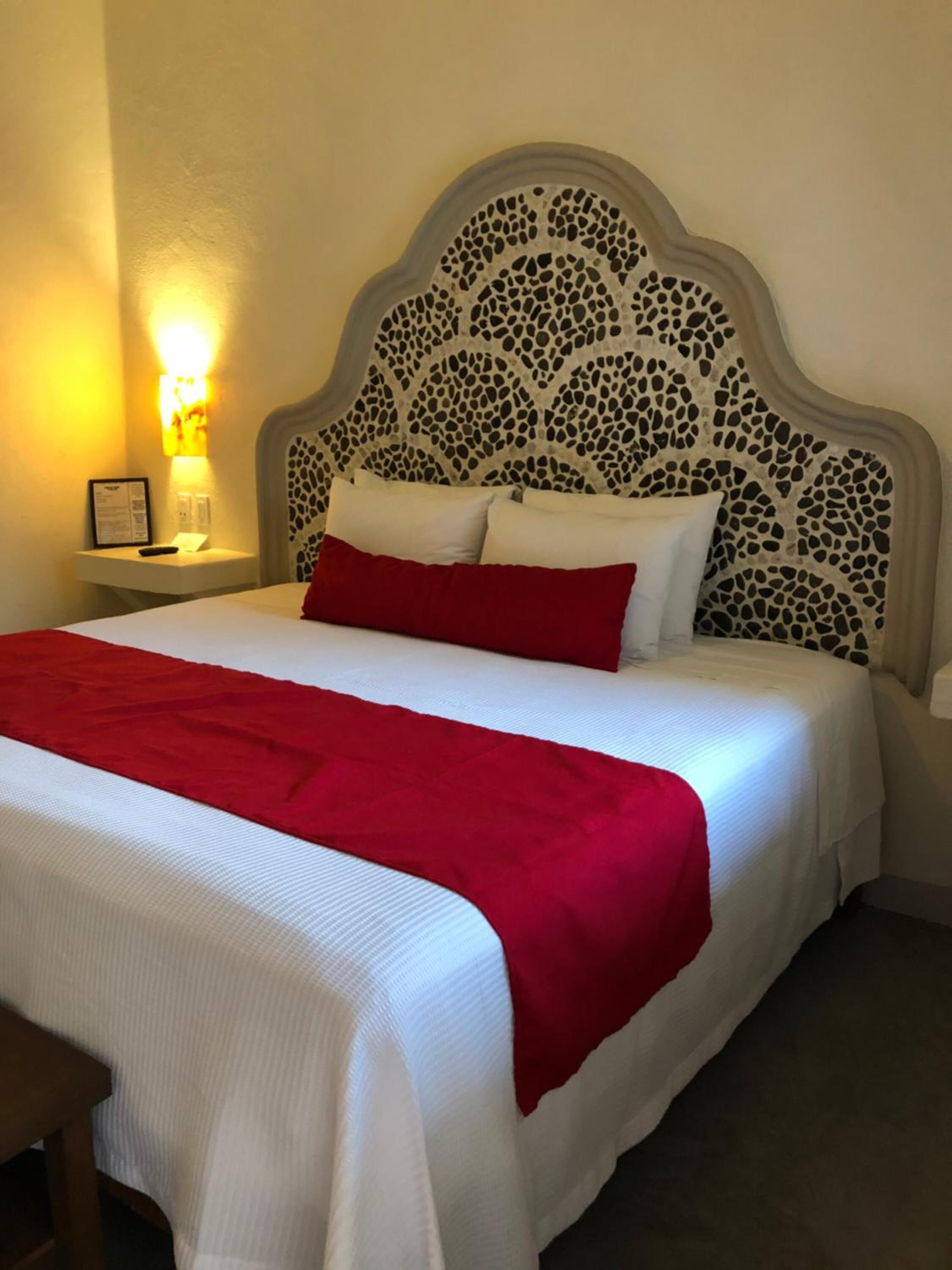 A neatly made bed with white and red bedding, a decorative headboard, and a bedside table with a lamp and a framed item.